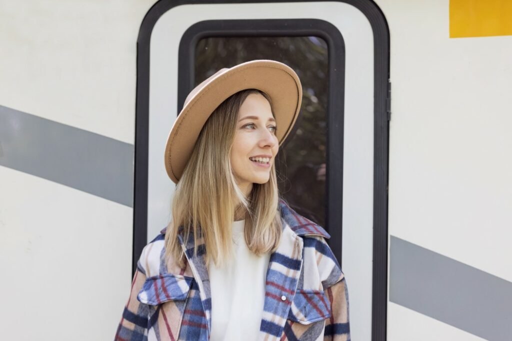 Una mujer con sombrero marrón y chaqueta azul a cuadros sale de su caravana y sonríe a su izquierda.