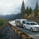 White Ford Expedition arrastra un remolque de viaje Airstream por una ladera cubierta de pinos.