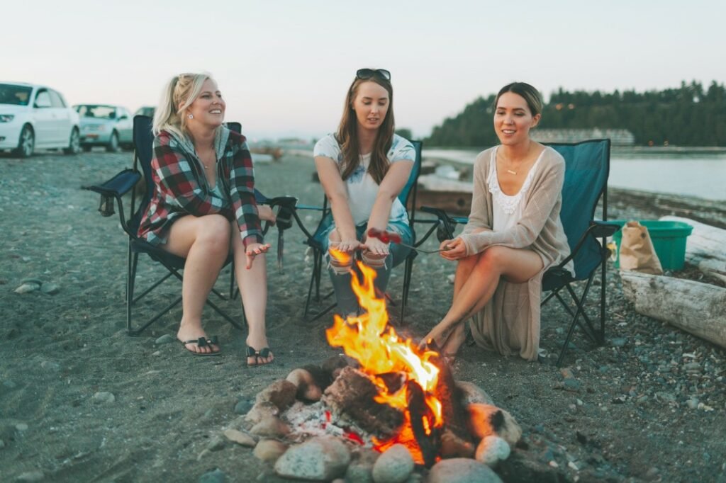Tres mujeres acampando en la playa cocinar perritos calientes sobre el fuego con palos de fogata