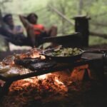 En primer plano, una cena de fogata de pollo, bistec y verduras se cocina en una sartén de hierro mientras dos campistas se relajan en una hamaca en el fondo.