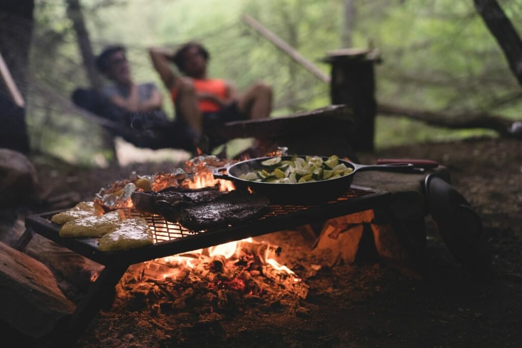 En primer plano, una cena de fogata de pollo, bistec y verduras se cocina en una sartén de hierro mientras dos campistas se relajan en una hamaca en el fondo.