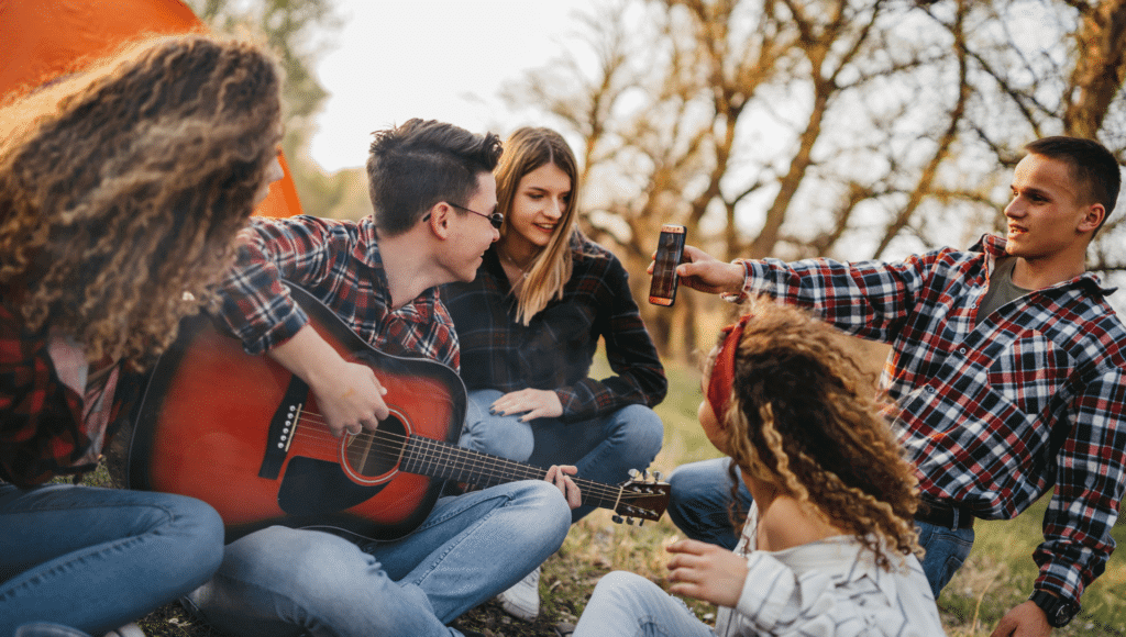 Un grupo de amigos se sienta en el suelo frente a una carpa.  Un hombre sostiene un teléfono mientras otro toca canciones de fogata en la guitarra.