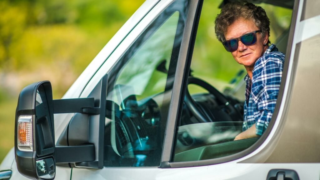 Una mujer con gafas de sol sentada en el asiento del conductor de una casa rodante mirando a la cámara por la ventana